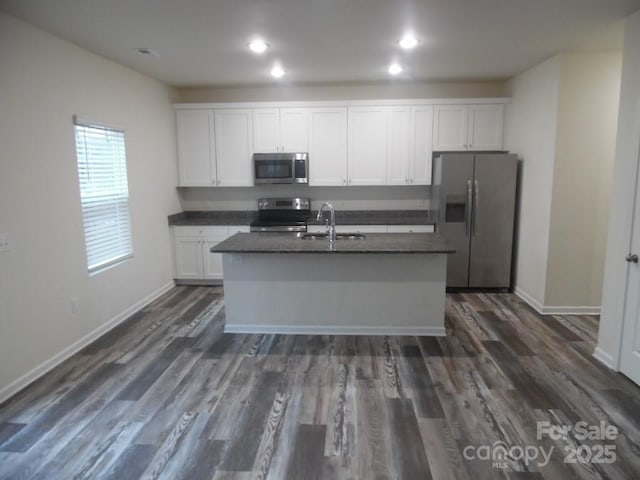 kitchen with stainless steel appliances, dark countertops, a center island with sink, and white cabinets