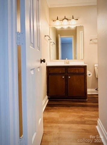 half bathroom with toilet, wood finished floors, vanity, baseboards, and crown molding