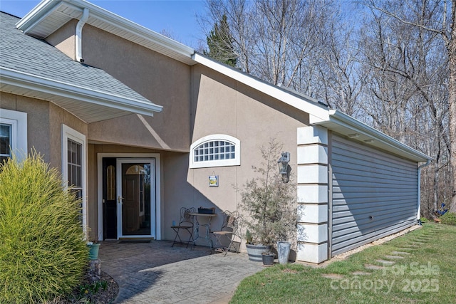 view of exterior entry featuring roof with shingles, a patio, and stucco siding