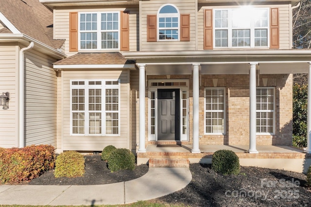 doorway to property featuring covered porch, brick siding, and roof with shingles