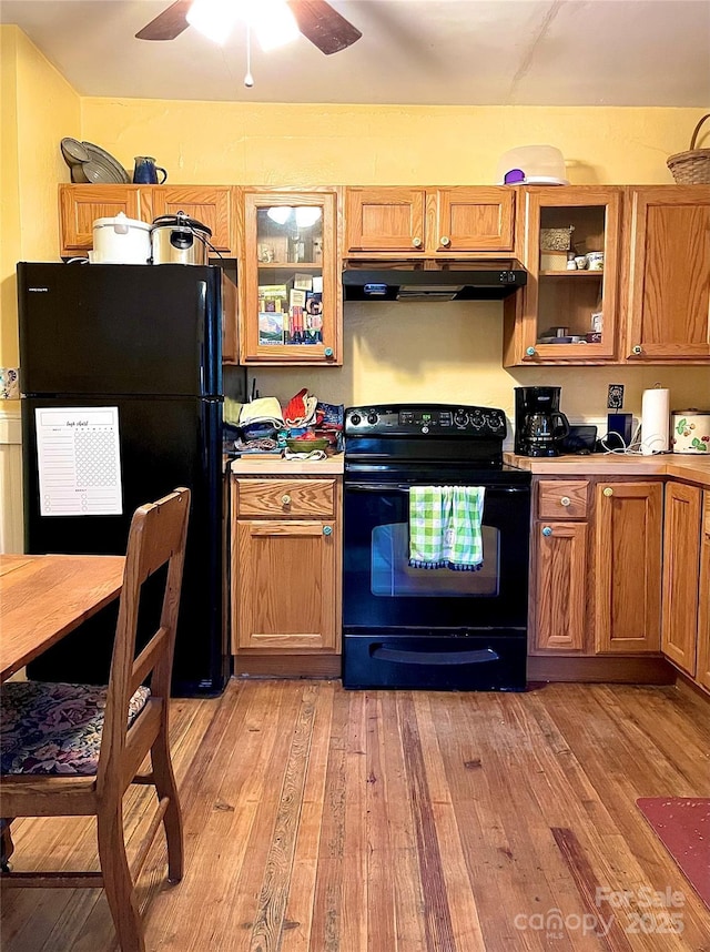 kitchen featuring light wood-type flooring, black appliances, under cabinet range hood, and glass insert cabinets