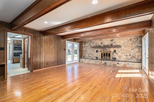 unfurnished living room with french doors, beam ceiling, a fireplace, wood walls, and light wood-type flooring