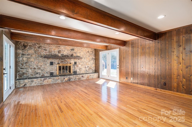 unfurnished living room with a stone fireplace, french doors, light wood-type flooring, wood walls, and beam ceiling