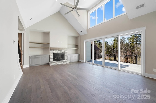 unfurnished living room with built in shelves, a stone fireplace, wood finished floors, visible vents, and a ceiling fan