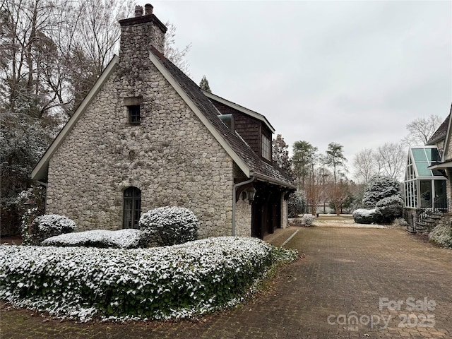 snow covered property featuring a garage, stone siding, a chimney, and driveway