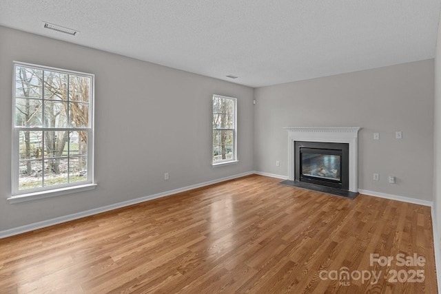 unfurnished living room with a textured ceiling, visible vents, baseboards, light wood finished floors, and a glass covered fireplace