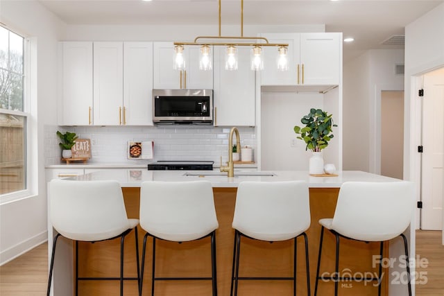 kitchen featuring white cabinets, stainless steel microwave, a kitchen island with sink, and decorative backsplash