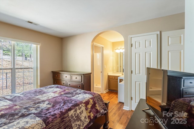 bedroom featuring visible vents, arched walkways, baseboards, ensuite bath, and light wood-type flooring