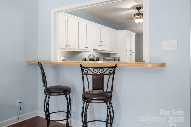 kitchen featuring under cabinet range hood, a breakfast bar, a ceiling fan, baseboards, and white cabinets