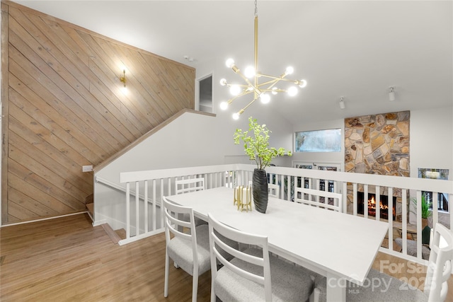 dining room featuring a notable chandelier, light wood finished floors, wood walls, vaulted ceiling, and a stone fireplace