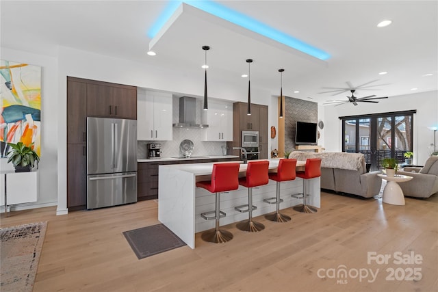 kitchen with wall chimney exhaust hood, appliances with stainless steel finishes, a breakfast bar, open floor plan, and light countertops