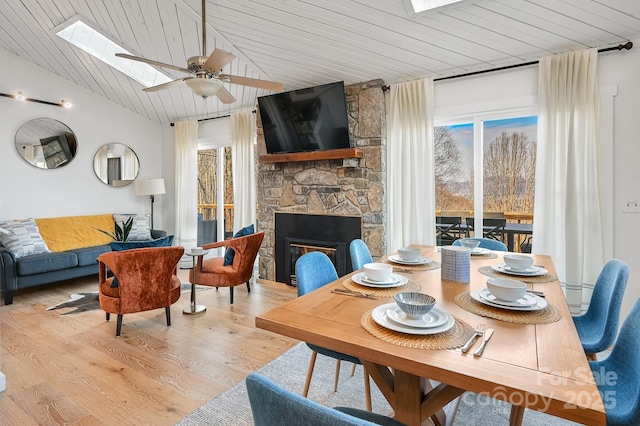 dining area with vaulted ceiling with skylight, a stone fireplace, light wood-style flooring, wood ceiling, and a ceiling fan