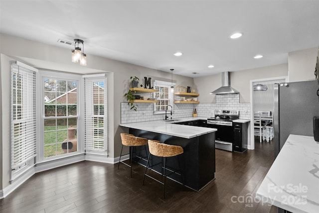 kitchen featuring wall chimney exhaust hood, a peninsula, stainless steel appliances, open shelves, and a sink