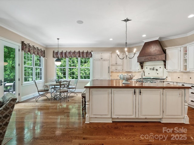 kitchen featuring custom range hood, hanging light fixtures, a kitchen island with sink, wooden counters, and backsplash