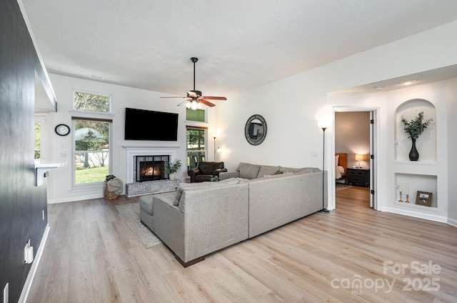 living area featuring a ceiling fan, a textured ceiling, light wood-type flooring, a lit fireplace, and baseboards