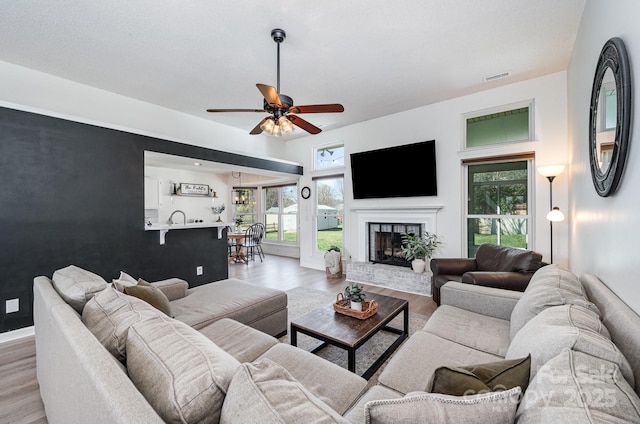 living room featuring ceiling fan, a fireplace, visible vents, baseboards, and light wood finished floors