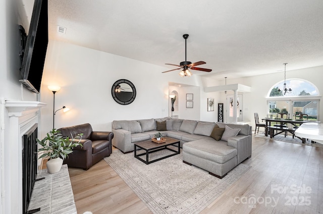 living area with visible vents, a textured ceiling, light wood-type flooring, a fireplace, and ceiling fan with notable chandelier