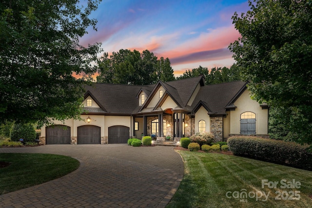 french country style house with decorative driveway, stone siding, a lawn, and stucco siding