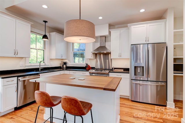 kitchen with stainless steel appliances, a sink, white cabinets, wall chimney range hood, and decorative light fixtures