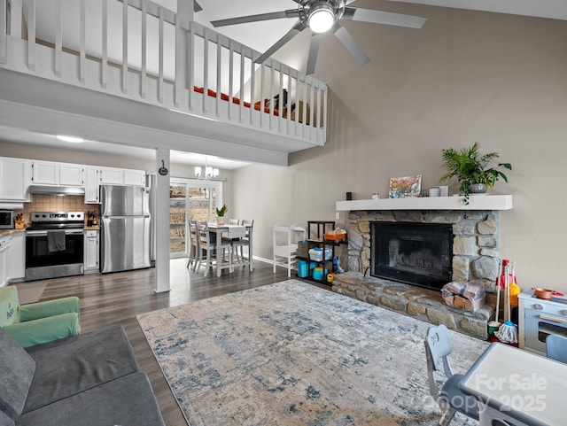 living room featuring ceiling fan with notable chandelier, dark wood-style floors, a stone fireplace, baseboards, and a towering ceiling