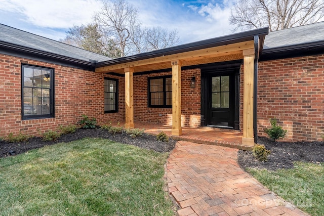 view of exterior entry with roof with shingles, a lawn, and brick siding