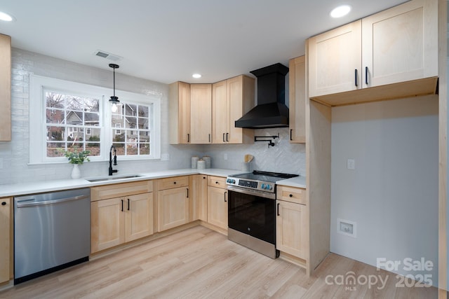 kitchen with light brown cabinets, a sink, appliances with stainless steel finishes, light wood-type flooring, and wall chimney exhaust hood