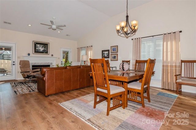 dining space with high vaulted ceiling, ceiling fan with notable chandelier, a high end fireplace, visible vents, and light wood-type flooring