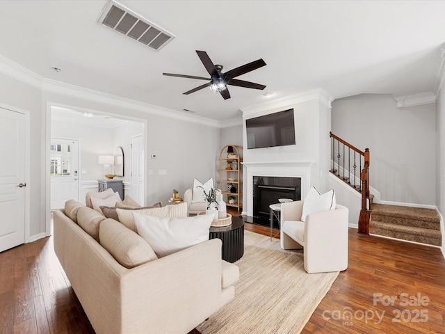 living area featuring a fireplace with flush hearth, visible vents, stairway, hardwood / wood-style floors, and crown molding