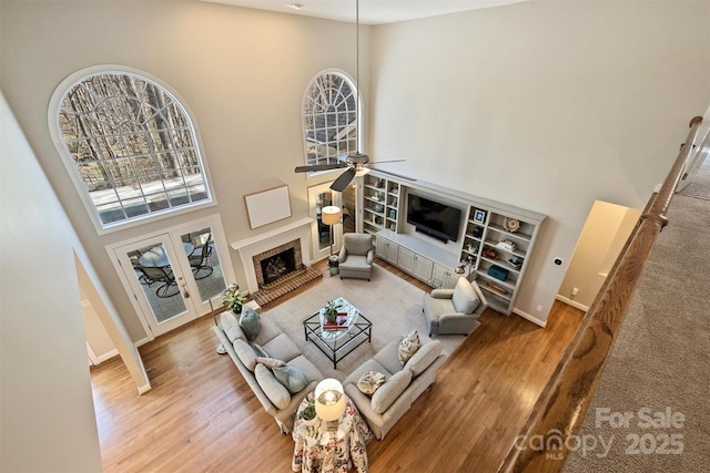 living area featuring a fireplace with raised hearth, a high ceiling, a ceiling fan, wood finished floors, and baseboards