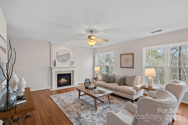 living room with ceiling fan, wood finished floors, a fireplace with flush hearth, visible vents, and baseboards