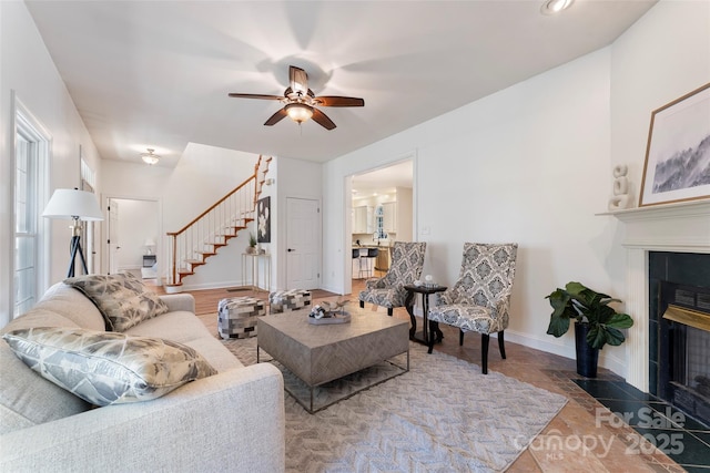 living room with ceiling fan, stairway, a tiled fireplace, and baseboards