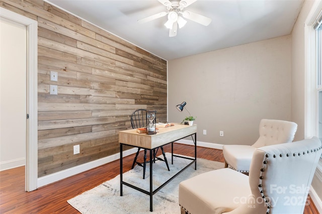 home office with an accent wall, ceiling fan, wood walls, wood finished floors, and baseboards