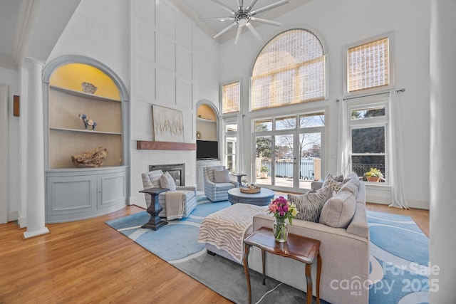 living area featuring a large fireplace, built in features, a ceiling fan, a towering ceiling, and wood finished floors