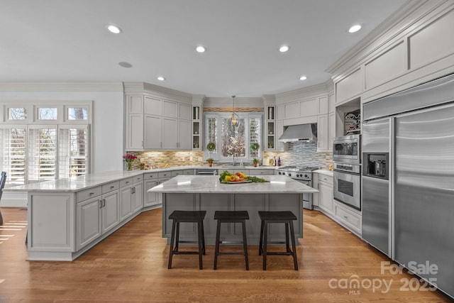 kitchen featuring built in appliances, a kitchen island, hanging light fixtures, wall chimney range hood, and glass insert cabinets