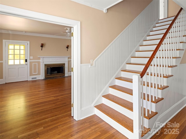 staircase with ceiling fan, a wainscoted wall, a fireplace with flush hearth, wood finished floors, and ornamental molding