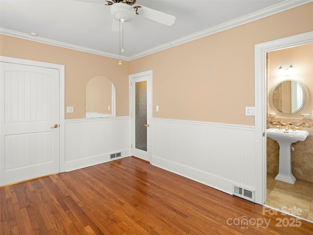 empty room featuring a wainscoted wall, wood finished floors, a ceiling fan, visible vents, and crown molding