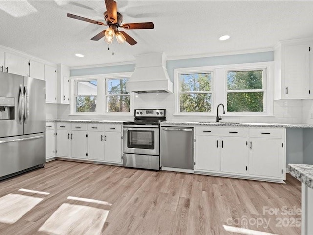 kitchen with light wood finished floors, stainless steel appliances, white cabinetry, a sink, and premium range hood