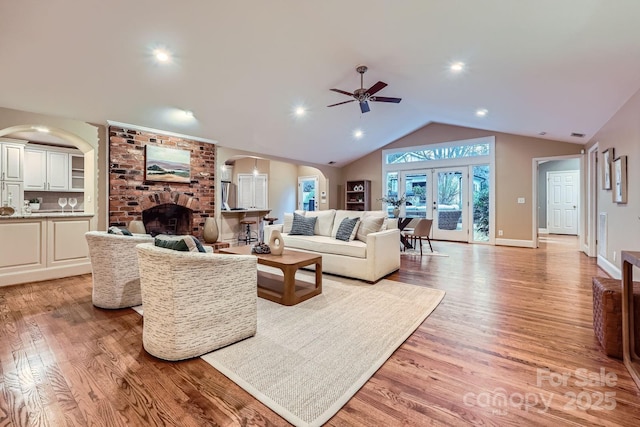 living room with arched walkways, ceiling fan, vaulted ceiling, a brick fireplace, and light wood-type flooring