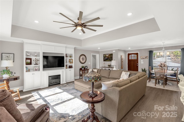 living area featuring a raised ceiling, a glass covered fireplace, wood finished floors, crown molding, and ceiling fan with notable chandelier