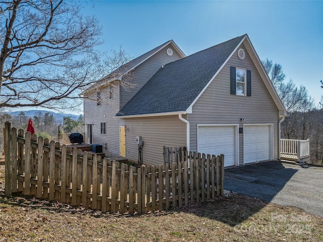 view of side of home featuring a garage, driveway, a shingled roof, and fence