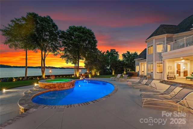 pool at dusk featuring a pool with connected hot tub, a patio area, a water view, and french doors