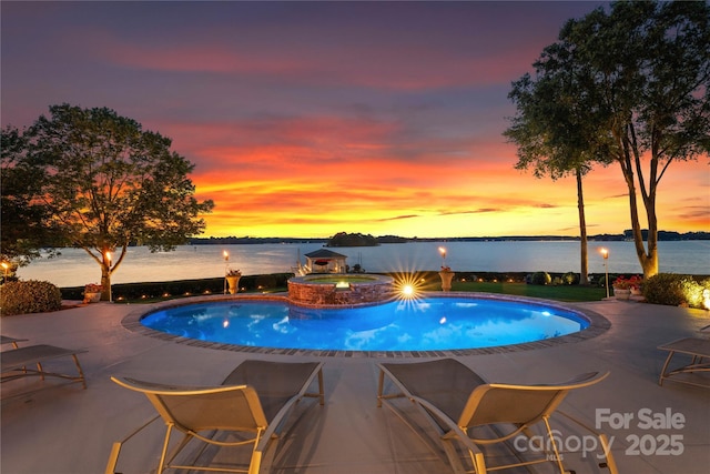 pool at dusk featuring a patio area, a pool with connected hot tub, and a water view