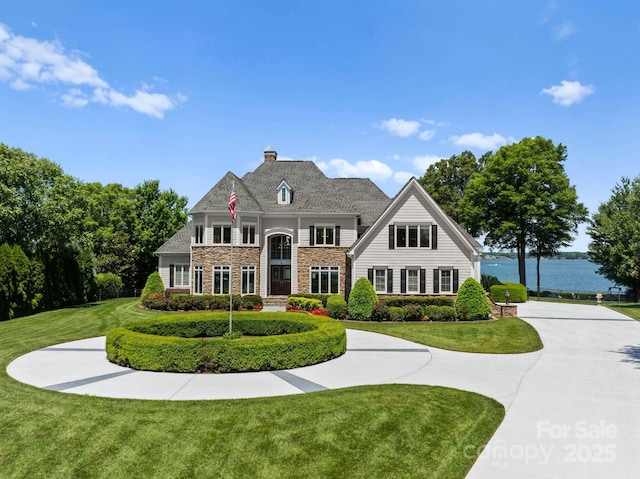 view of front of property featuring a front yard, a chimney, curved driveway, stone siding, and a water view