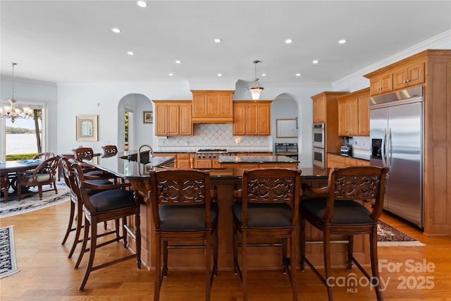 kitchen featuring light wood-style flooring, dark countertops, arched walkways, appliances with stainless steel finishes, and a large island with sink