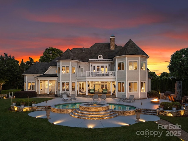 back of house at dusk featuring a pool with connected hot tub, a fire pit, a lawn, a balcony, and a patio area