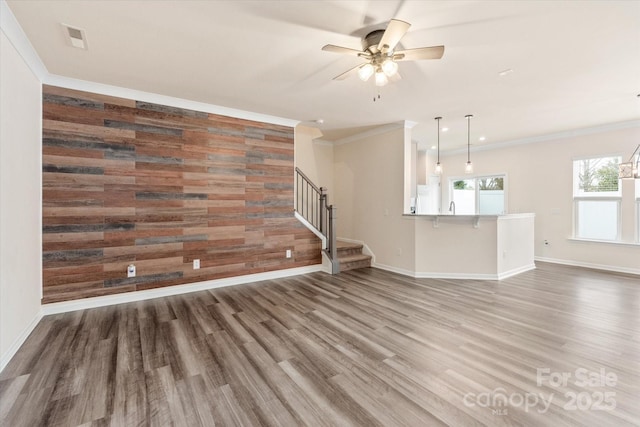 unfurnished living room with an accent wall, wood walls, wood finished floors, ornamental molding, and stairway