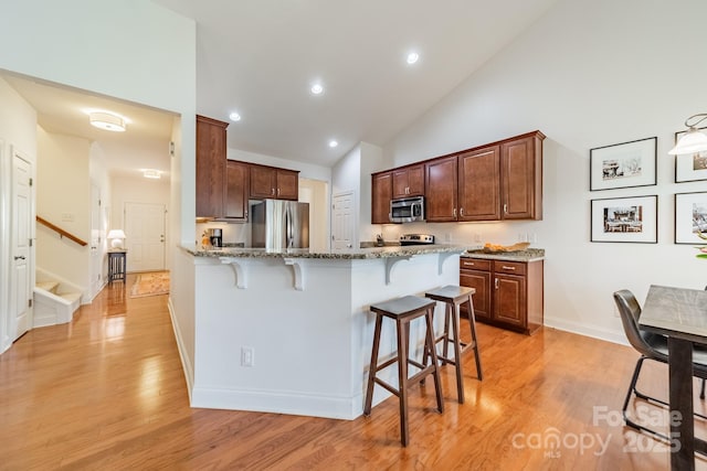 kitchen featuring appliances with stainless steel finishes, light stone countertops, light wood-type flooring, a kitchen bar, and high vaulted ceiling