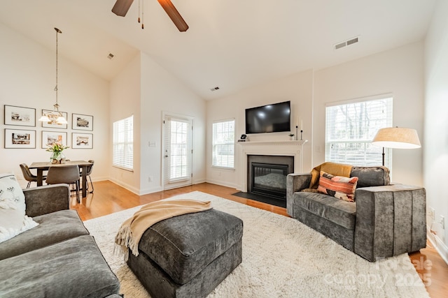 living area featuring visible vents, light wood-style flooring, a fireplace with flush hearth, high vaulted ceiling, and baseboards