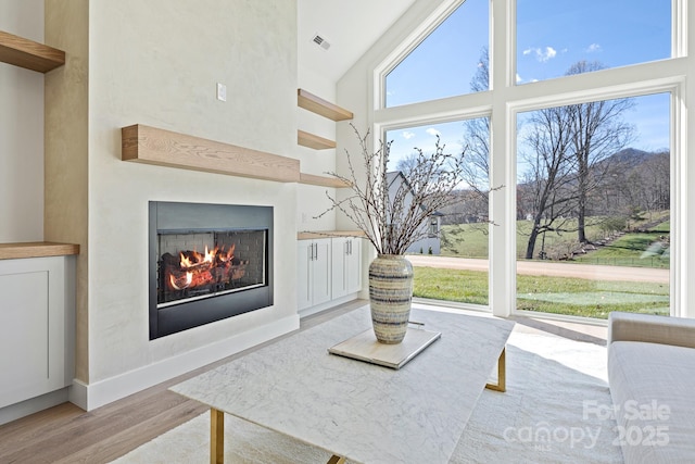 living area featuring visible vents, baseboards, light wood-style flooring, a lit fireplace, and a high ceiling