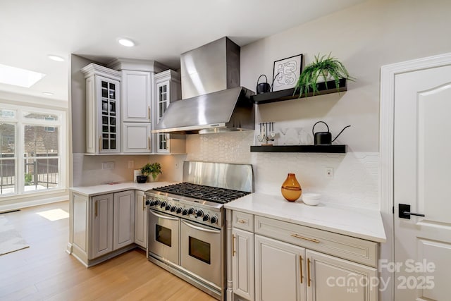 kitchen with a skylight, range with two ovens, wall chimney exhaust hood, glass insert cabinets, and backsplash